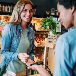A woman in a produce market holds a credit card above a cashier's hand, who holds a payment terminal at checkout.