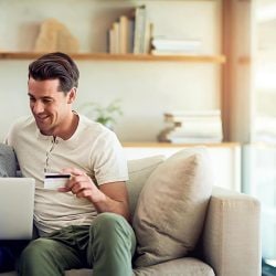 A couple sits together on a couch looking at a laptop. The man holds a credit card in his hand.