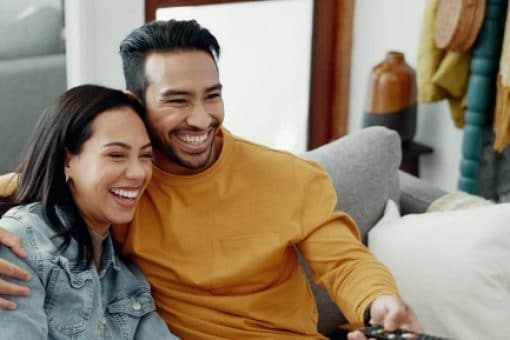 A smiling couple sit closely together on a couch. The man holds a TV remote.