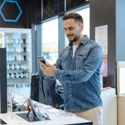 A man tests a smartphone for sale in a phone store, surrounded by displays of smart phones and other devices.