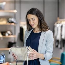 Smiling woman shopping in a boutique holding a brown handbag, surrounded by stylish clothing and accessories in a modern store setting.