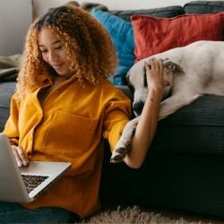 A smiling woman sits on the floor in front of a couch, working on her laptop and petting a dog.