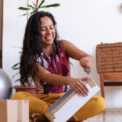 A woman sits on the floor smiling as she uses a tool to open boxes in her apartment.