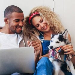 A young couple looks over documents on their laptop while also playing with their husky puppy.