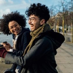 A man and a woman holding coffee to-go cups laugh while looking out over a river.