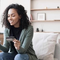 A woman sits on a chair looking at her mobile phone and holding a credit card in her other hand.