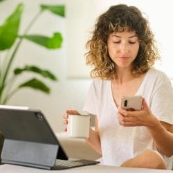 A woman sits on a couch holding a coffee mug and looking at her mobile phone.