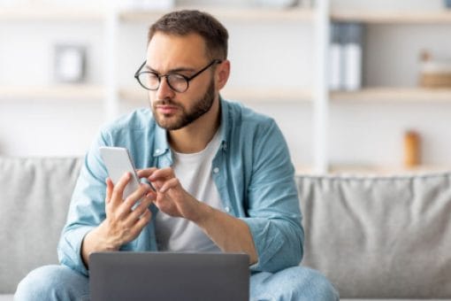 A man sits on a couch, scrolling on his phone in front of an open laptop.