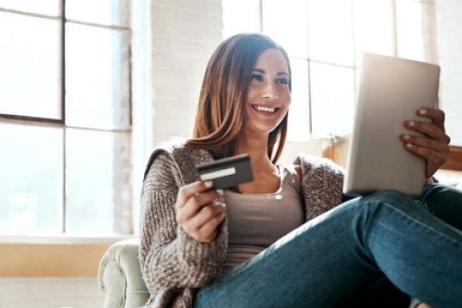A woman sits on a couch holding a credit card and reviewing her credit score on a tablet.