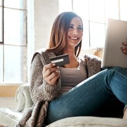 A woman sits on a couch holding a credit card and reviewing her credit score on a tablet.