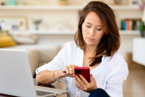 A woman scrolls on her phone and sits in front of a laptop reviewing her credit.