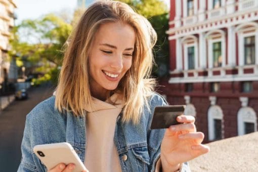 A woman stands outside holding her phone in one hand and a credit card in the other.