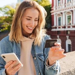 A woman stands outside holding her phone in one hand and a credit card in the other.