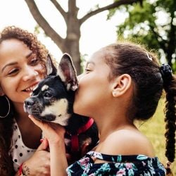 A young girl kisses her pet as her mom watches and smiles.