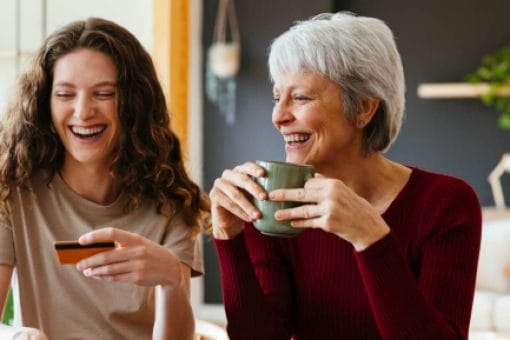 An older woman and a younger woman smile and laugh together. The older woman holds a mug. The younger woman holds a credit card and pushes keys on a laptop.