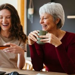 An older woman and a younger woman smile and laugh together. The older woman holds a mug. The younger woman holds a credit card and pushes keys on a laptop.