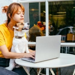 Woman sits outside with her dog in lap typing on her laptop