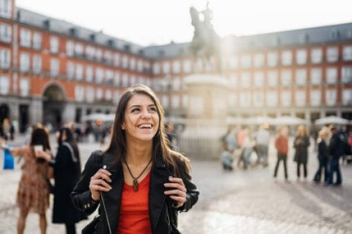 A woman visiting another country walks through a courtyard with a large statue and groups of people.