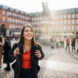 A woman visiting another country walks through a courtyard with a large statue and groups of people.