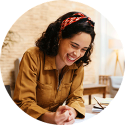 Smiling woman leans over desk with hands clasped next to a stack of notepads.