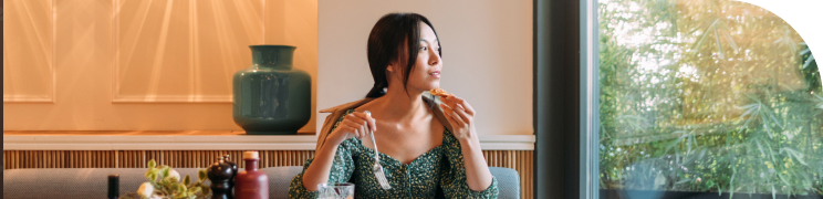 A woman sits at a table looking outside through a bright window. She has a fork in one hand and a bite of food in the other.