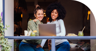 Two women sit together in an open cafe window. They both look at a laptop in front of them.