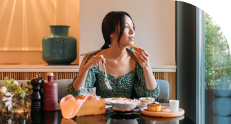 A woman sits at a table looking outside through a bright window. She has a fork in one hand and a bite of food in the other.