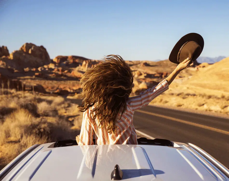 woman stands up through car sunroof waving her hat with a rocky desert landscape around her