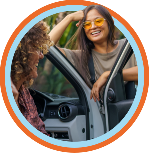 Young woman with orange sunglasses leans on an open car door while taking to her friend