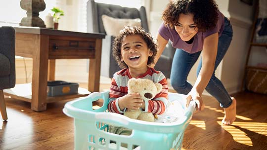 A little boy giggles as a young woman pushes him around the living room floor in a laundry basket.