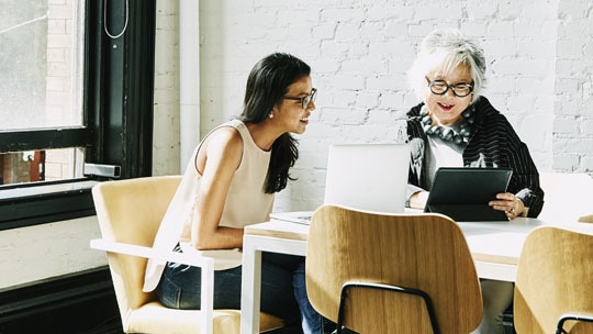 Two women in conversation while looking at laptops.