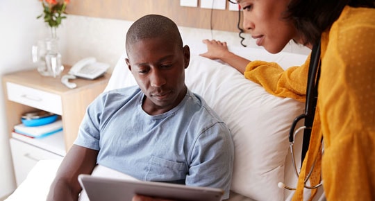 Man in hospital bed talking to staff member while looking at computer tablet.