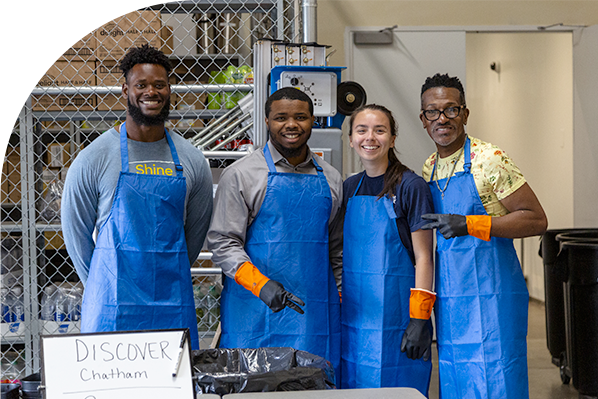 Four Discover employees smiling and standing together in a warehouse, wearing matching blue aprons and orange arm bands. 