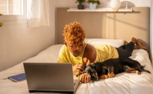 A woman using her laptop while reclining in bed with her sleeping dog.