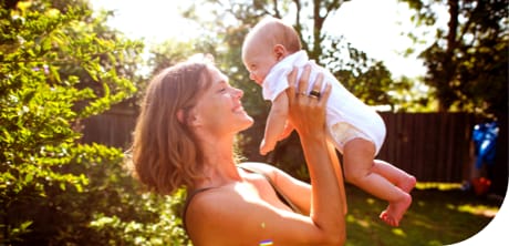 A woman holds an infant in the air playing in a fenced-in backyard
