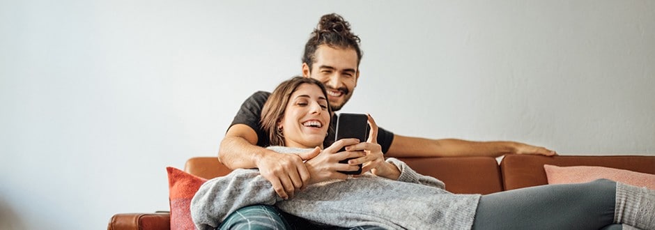 Smiling young couple using smartphone sitting on sofa at home