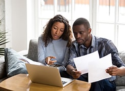 Serious African American couple discussing paper documents, sitting together on couch at home, man and woman checking bills, bank account balance, terms of contract, mortgage, loan agreement