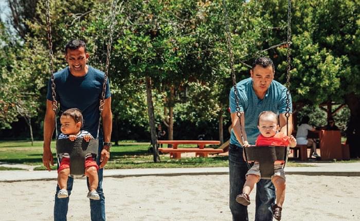 Two men are pushing kids on swings in a park.