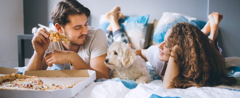 A couple laying on their bed, with their dog, and eating pizza out of the box.