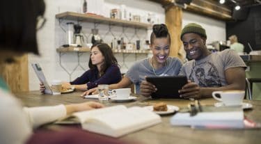 A group of people in an office breakroom. One reads and two look at a phone.