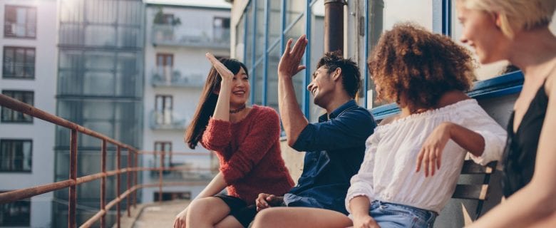 Four friends sitting on the balcony of a city apartment building, two of them giving each other a high-five.