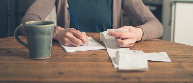 Closeup of a man writing down information while looking at a long receipt and drinking a cup of coffee.