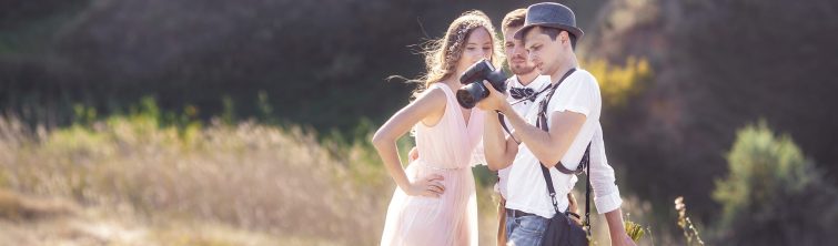 A photographer shows an engaged couple a picture on the screen of his camera. They are standing in a field.