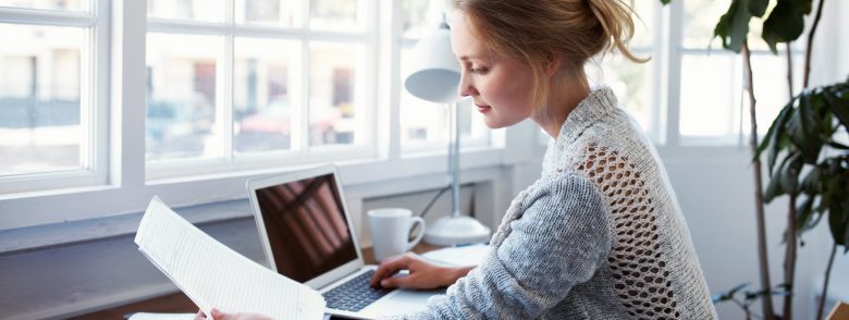 A woman sits at her home desk, with a laptop computer in front of her, while reading some papers.