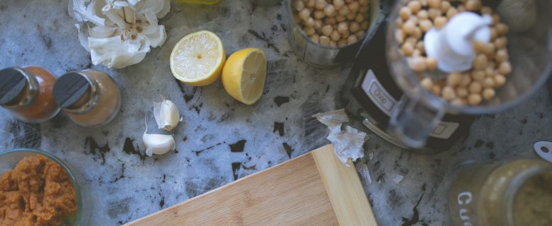 A counter top with a cutting board, food processor, and various ingredients for making hummus.