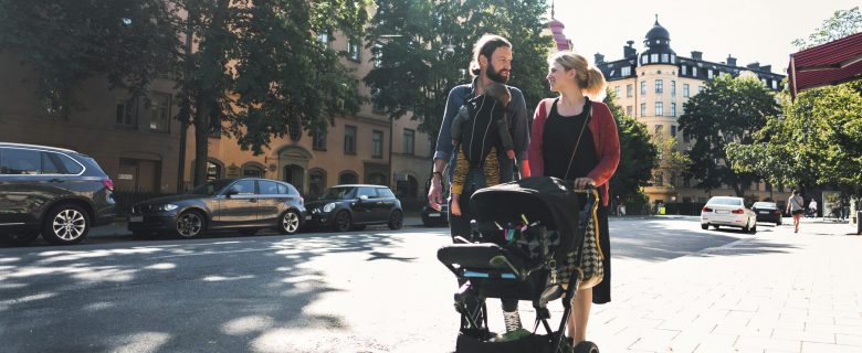 Parents smile at each other walking down the street. The father has a baby strapped to his chest while the mother pushes a second baby in a stroller.