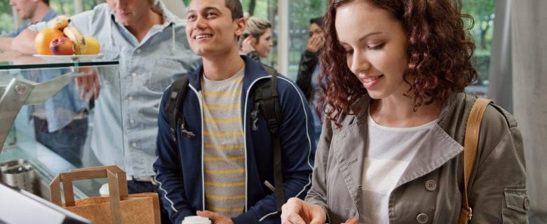 College students are lined up at a dining hall cafeteria. A college-age woman and man are smiling at the front of the line. 