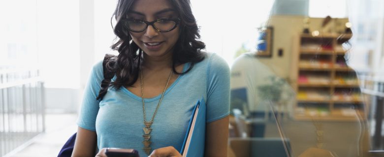 A college woman with a backpack on one shoulder and a binder underneath her arm checks her phone as she leaves a classroom. 