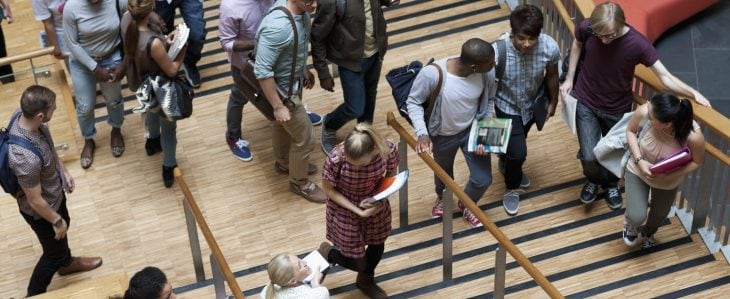 A group of college students holding books in their hands and wearing bags and backpacks walk up a staircase. 