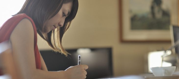 A woman, sitting down, takes notes with a cup of coffee in front of her.